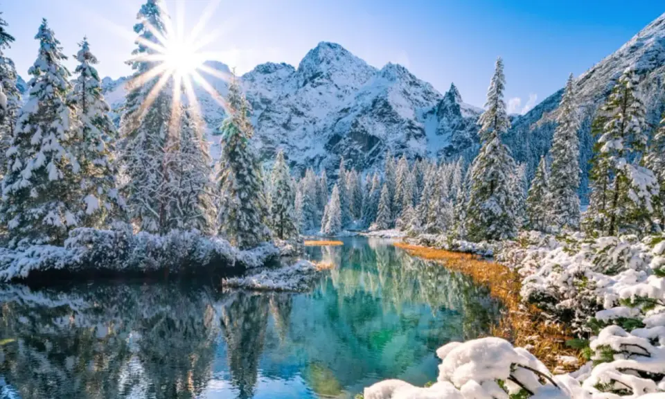 Morskie Oko zimą. Słońce przebija się przez ośnieżone drzewa, odbijając się w turkusowej tafli wody. W tle majestatyczne, pokryte śniegiem góry.