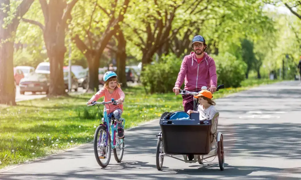 Radosne dziecko na rowerze jedzie przed rodzicem, który wiezie drugie dziecko w przyczepce rowerowej.