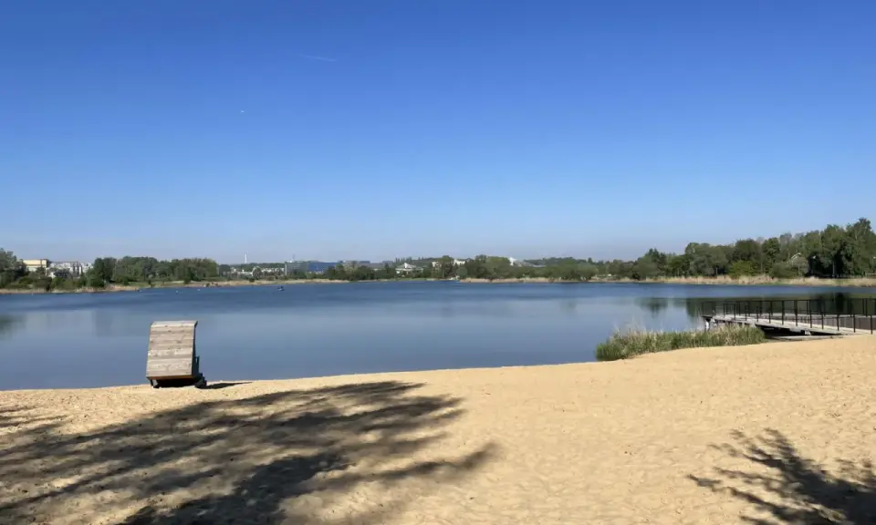 Plaża bagry z drewnianą przebieralnią i pomostem, w tle jezioro i zieleń.