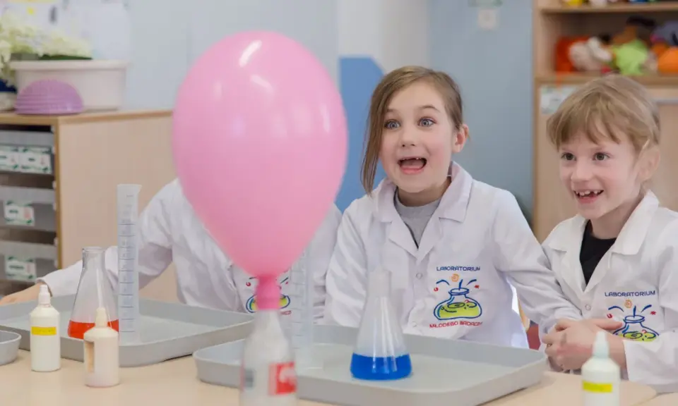 Dzieci w fartuchach laboratoryjnych z ekscytacją obserwują dmuchany balon podczas eksperymentu. To świetna chemia dla dzieci!