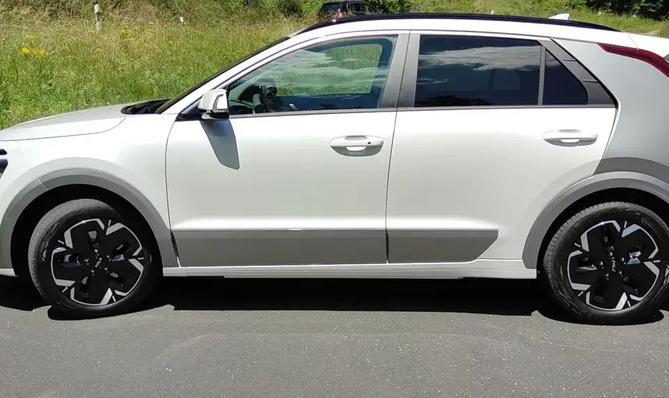 Biały Kia Niro EV z czarnymi felgami stoi na drodze, otoczony zielenią.