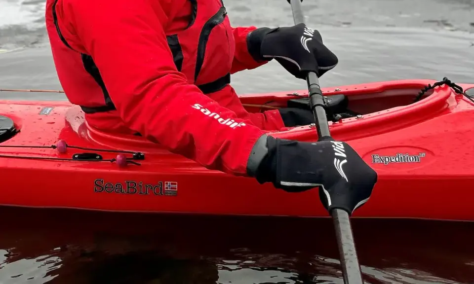 Person i röd jacka och svarta handskar paddlar kajak i kallt vatten.