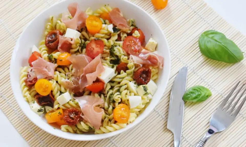 Une salade de pâtes originale avec des tomates cerises, du prosciutto, des dés de mozzarella et une sauce verte savoureuse.