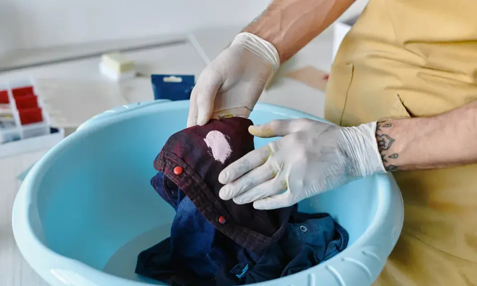 Hände mit Handschuhen behandeln ein dunkles Hemd mit einem hellen Fleck, um unbekannte Flecken zu entfernen.