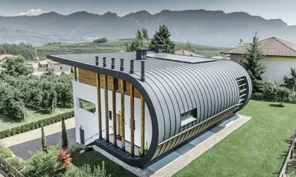 Nowoczesny dom z zaokrąglonym dachem z aluminium, które nie koroduje, otoczony zielenią i górami.