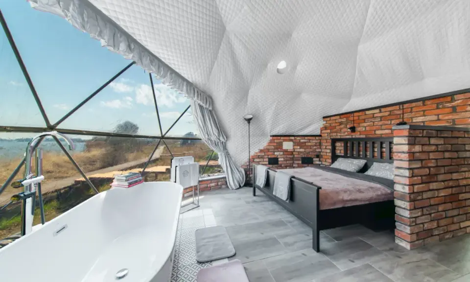 Luksusowy glamping nad jeziorem: wanna z widokiem, łóżko i ceglana ściana w kopułowym wnętrzu.