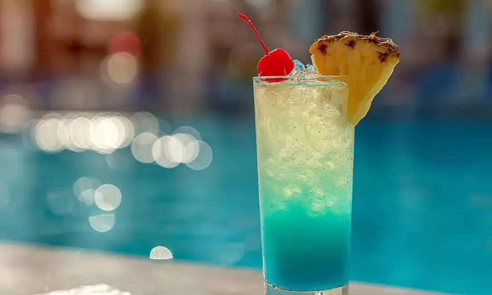 En blå och gul drink med körsbär och ananas står vid en pool. Perfekt för en solig dag.