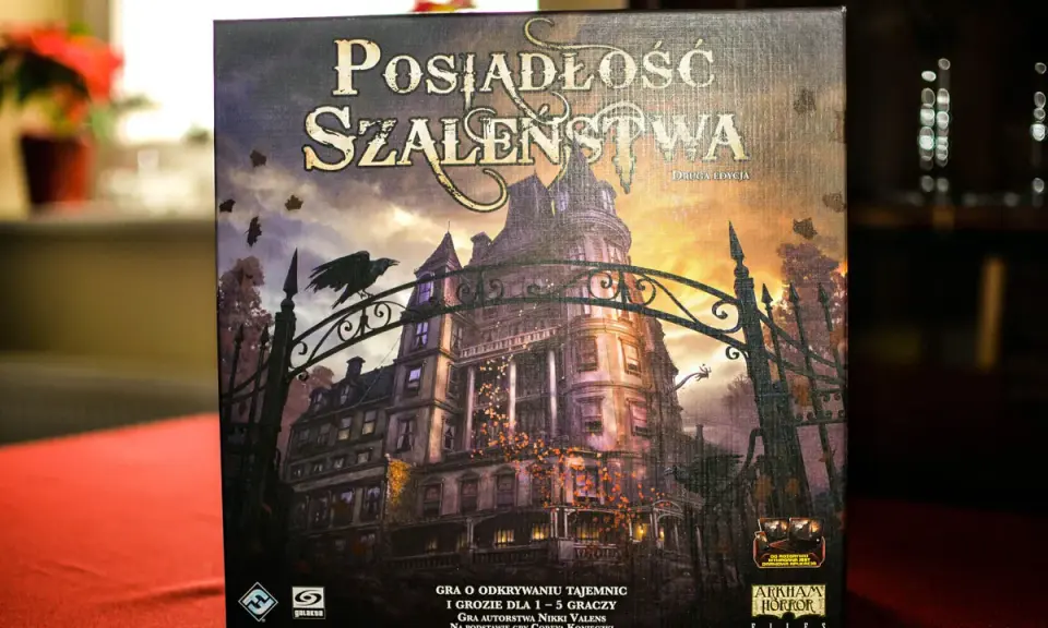 Pudełko gry "Posiadłość Szaleństwa" z mroczną posiadłością i wroną na bramie. Dodatki do tej gry planszowej zapowiadają nowe, przerażające przygody.