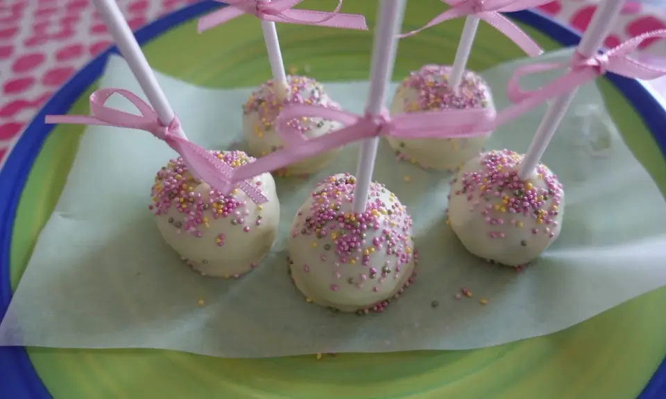 Sju söta cake pops med vit choklad och rosa, gula och lila strössel, dekorerade med rosa band.