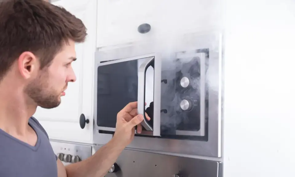 Mann öffnet Mikrowelle, aus der Rauch dringt. Ein Fehler mit Metall in Mikrowelle kann gefährlich sein.