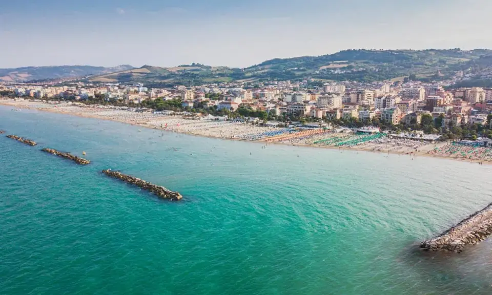 Vista aerea di San Benedetto del Tronto: una lunga spiaggia con ombrelloni colorati e il mare turchese, con dighe frangiflutti che proteggono la costa.