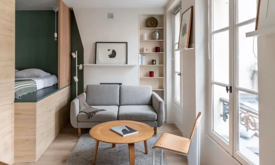 Aménagement petit appartement : lit surélevé, canapé gris, table basse ronde en bois, étagères et grande fenêtre lumineuse.