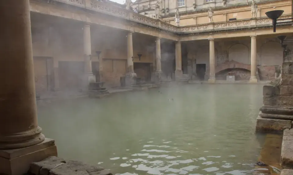 Dampf steigt aus dem Becken der römischen Therme auf. Säulen und Bögen umgeben das Wasser, in dem Enten schwimmen.