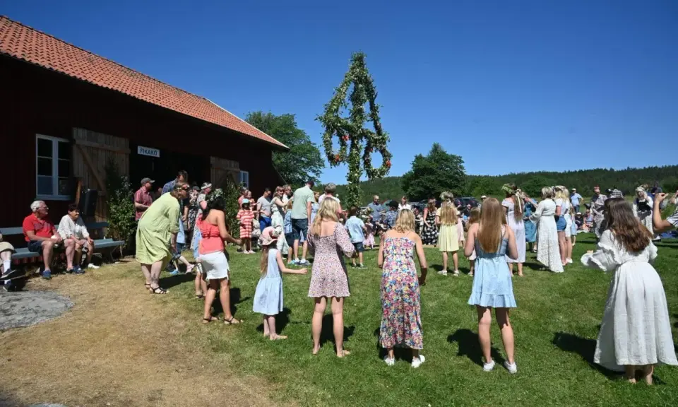 Folk dansar runt midsommarstången på midsommar afton. En vacker sommardag med klarblå himmel.