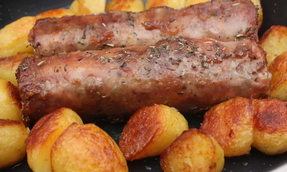 Deux andouillettes artisanales dorées, parsemées d'herbes, mijotent avec des pommes de terre rôties dans une poêle.