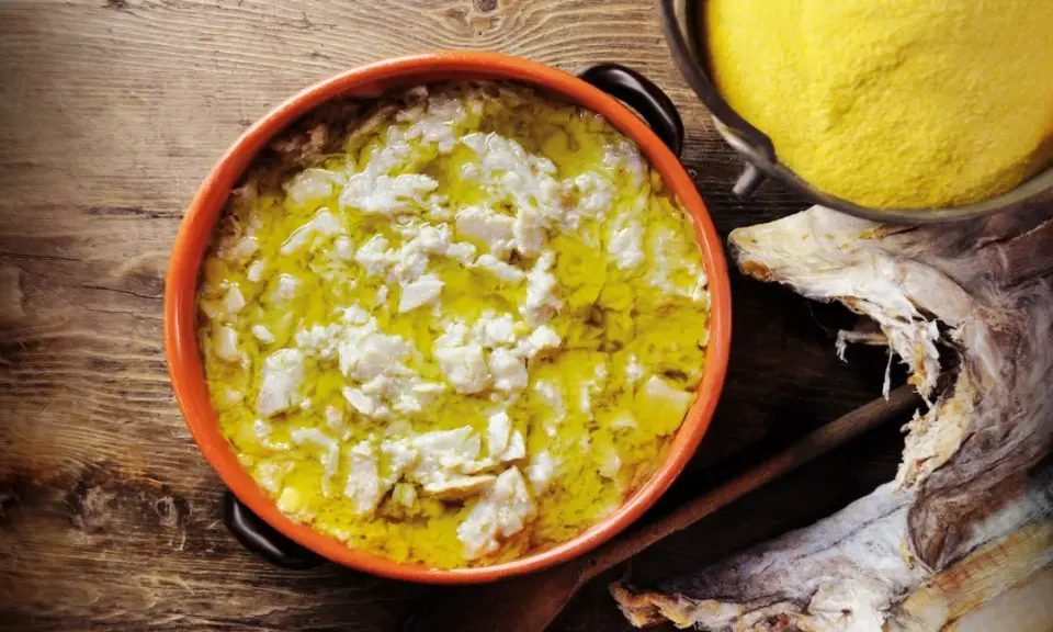 Piatto fumante di baccalà alla vicentina, con polenta gialla e stoccafisso essiccato sullo sfondo.