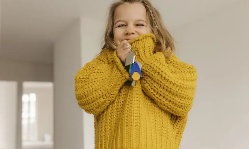 Ein fröhliches kleines Mädchen im gelben Pullover hält Schlüssel in der Hand. Die Frage "ab wann Kinder alleine lassen" beschäftigt Eltern oft, doch dieses Bild strahlt Unbeschwertheit aus.