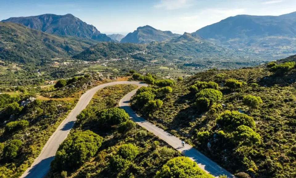 Ein Rennradfahrer auf Kreta genießt die kurvenreiche Straße durch eine grüne Berglandschaft.