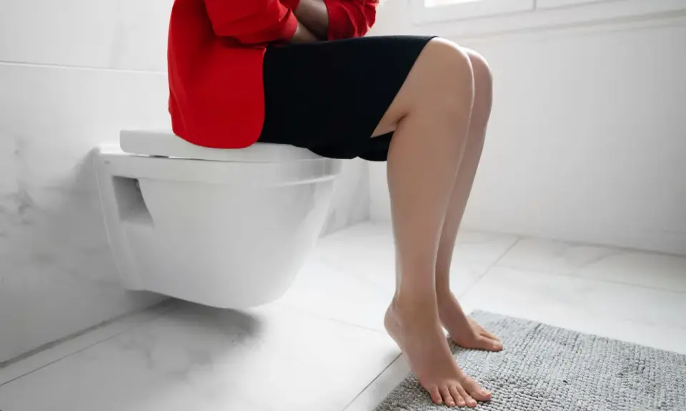 Kobieta w czerwonej marynarce i czarnej sukience siedzi na toalecie, trzymając się za brzuch. Szuka domowych sposobów na skąpomocz.