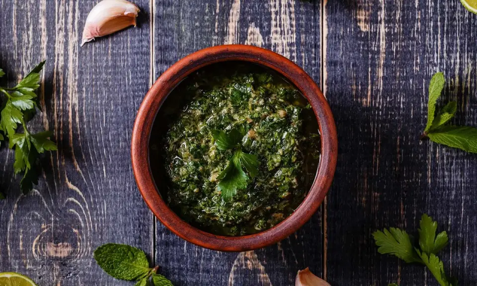 Un ciotola di terracotta contiene un pesto verde brillante, perfetto per accompagnare le acciughe al verde. Ingredienti freschi come aglio e prezzemolo circondano la ciotola.