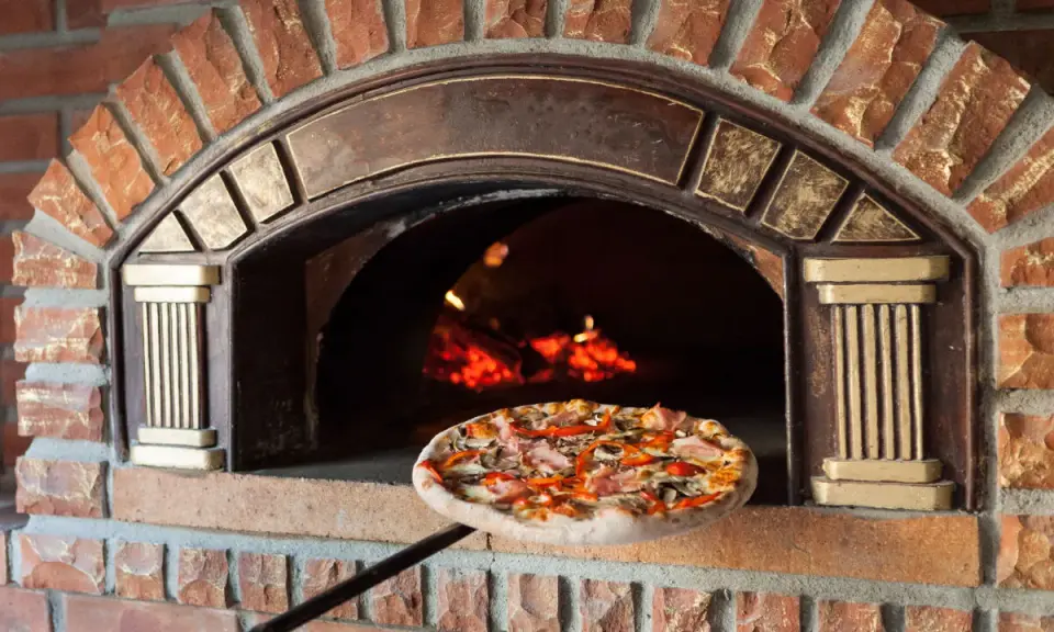 Une pizza garnie est sur le point d'entrer dans un four à pizza extérieur en briques rouges, avec des flammes visibles à l'intérieur.