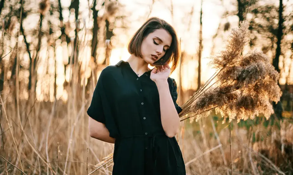 Młoda kobieta w czarnej sukience pozuje na tle zachodzącego słońca i trzcin. Idealna modelka do nauki kurs fotografii portretowej.
