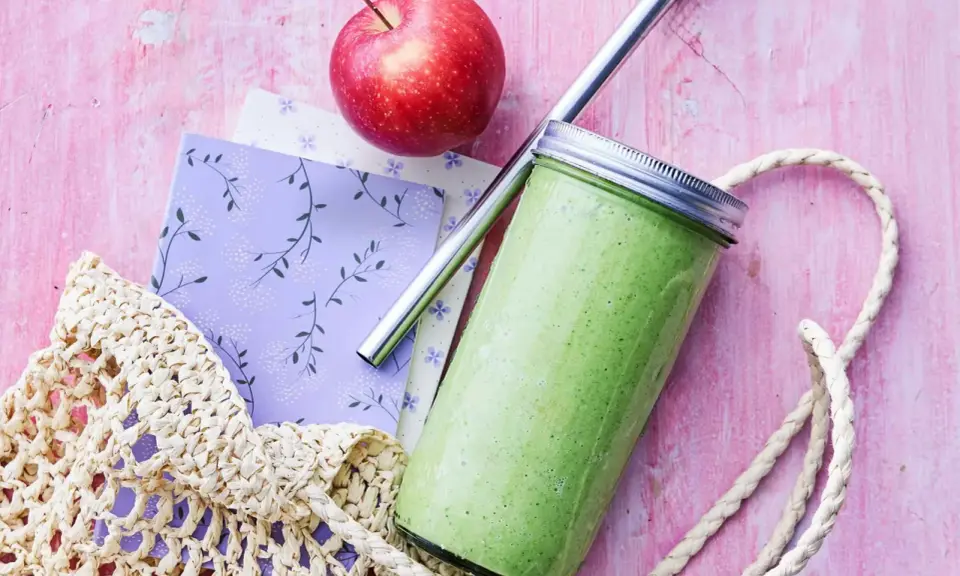 En grön, proteinrik smoothie i glasburk med metallrör, bredvid ett rött äpple och en vävd kasse på en rosa bakgrund.