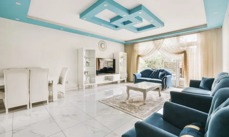 Nowoczesny salon z błękitnym, geometrycznym, dekoracyjnym malowidłem na suficie. Wygodne, granatowe sofy i białe meble tworzą elegancką przestrzeń.