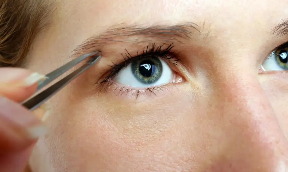 Une main utilise une pince à épiler pour affiner des sourcils, créant une ligne élégante au-dessus d'un œil bleu-vert.