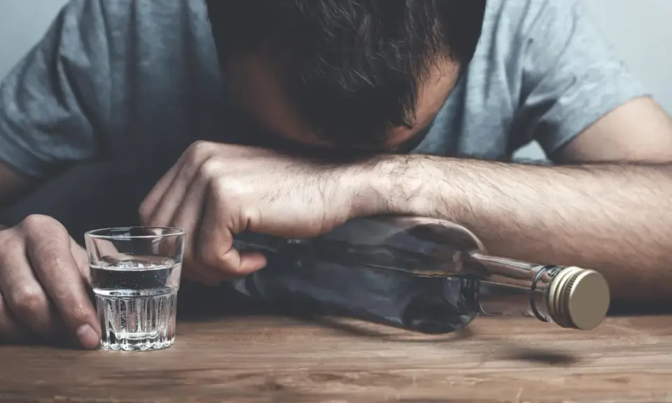 Mężczyzna z głową opartą o butelkę, obok kieliszek z alkoholem. Wygląda na to, że doświadcza zatrucia alkoholowego.