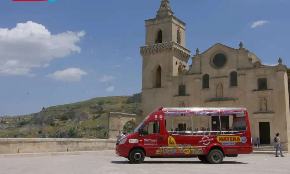 Un bus rosso "open top" per un tour di Matera, con la chiesa e i Sassi sullo sfondo.