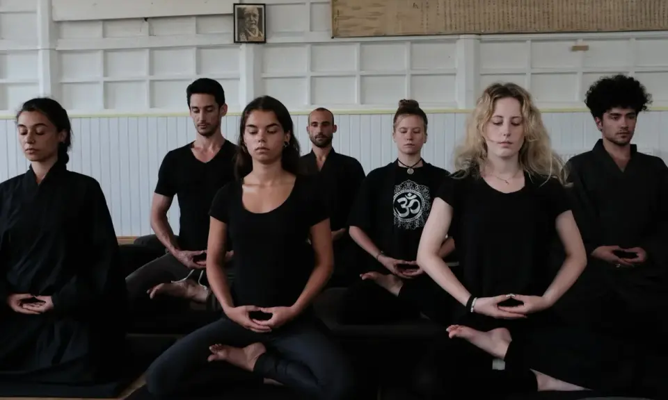 En grupp människor sitter i meditation, klädda i svarta kläder. En zen master leder sessionen.
