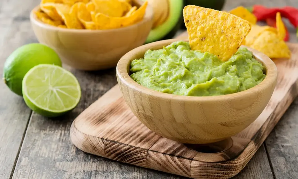 Miseczka guacamole z nachos, limonką i awokado na drewnianym tle.