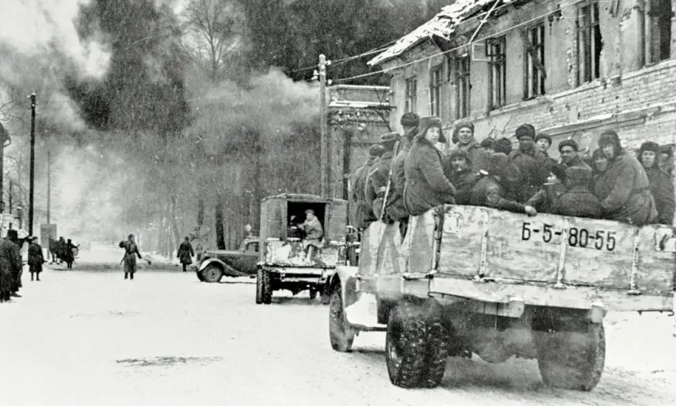 Soldaten der Roten Armee 1945 fahren in einem LKW durch eine verschneite Stadt. Rauch steigt aus einem zerstörten Gebäude auf.
