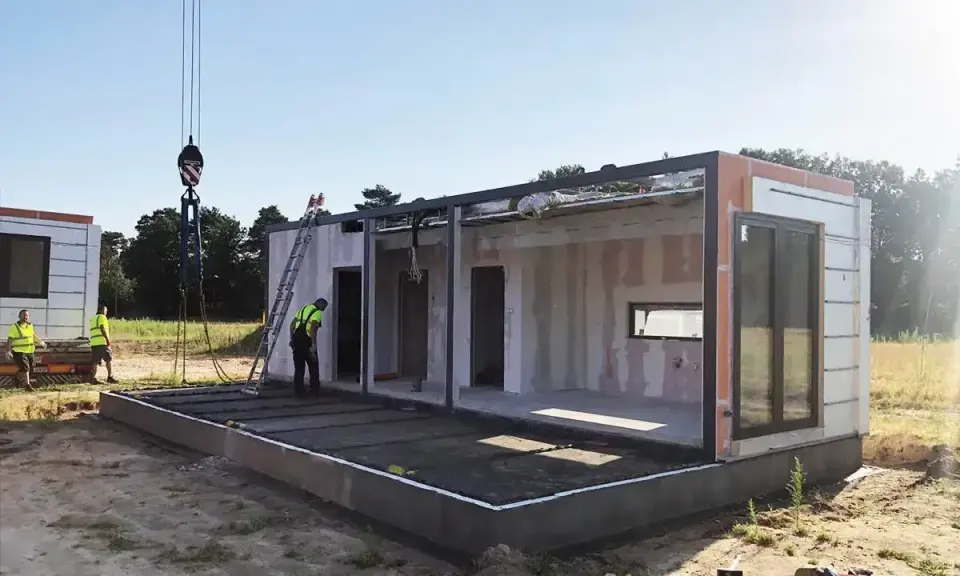 Budowa domu na zgłoszenie postępuje. Dźwig podnosi moduł, a pracownicy w kamizelkach nadzorują montaż. Warunki zabudowy sprzyjają szybkiemu postawieniu konstrukcji.