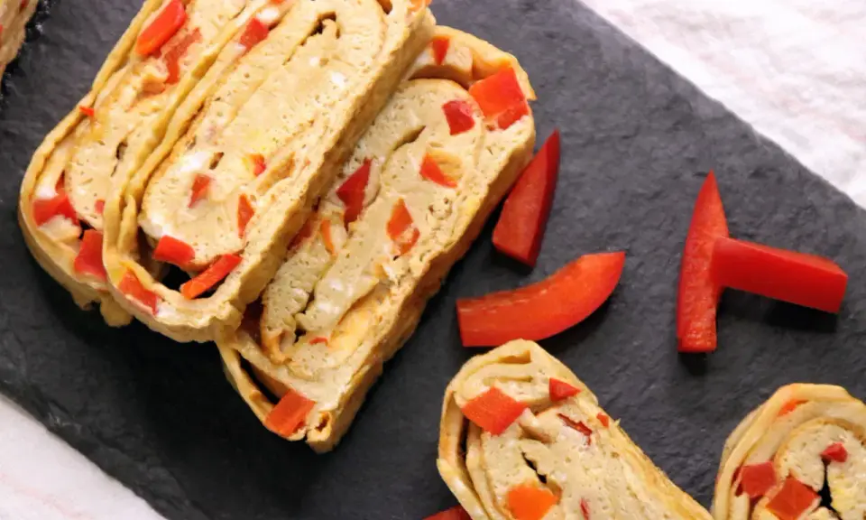 Roladki omletu tamago z kawałkami czerwonej papryki, ułożone na czarnej desce.