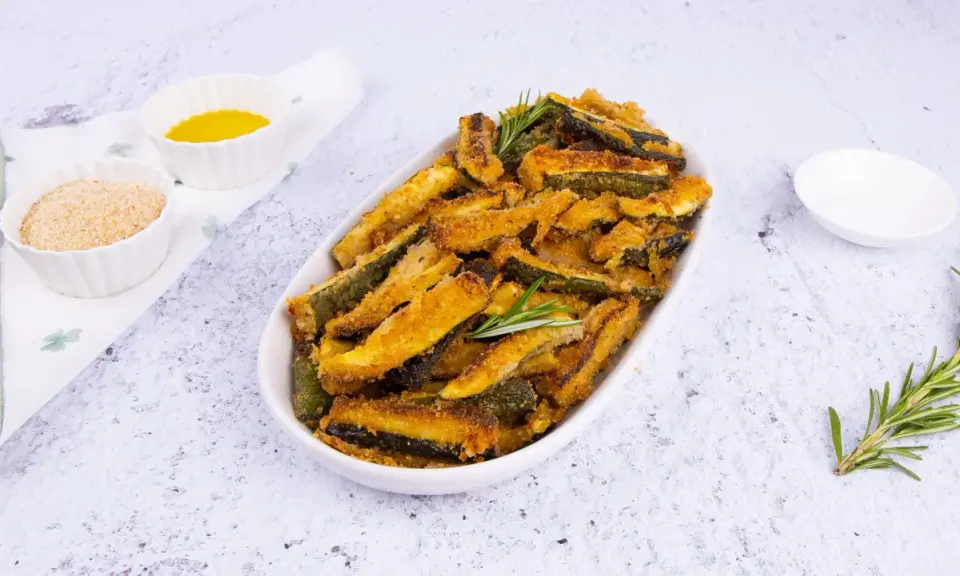 Zucchine gratinate al forno, dorate e croccanti, servite in un piatto ovale con rosmarino fresco. Accanto, ciotoline con pangrattato e olio.