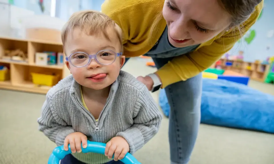 Chłopiec z zespołem Downa, w okularach, z językiem na brodzie, bawi się z terapeutką. To przykład pracy w obszarze oligofrenopedagogiki.