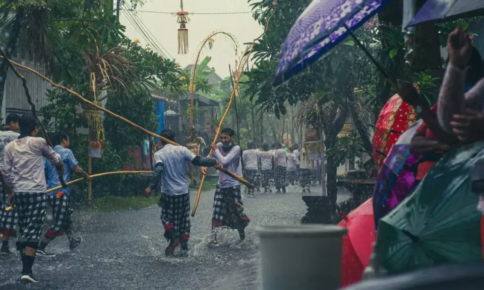 Ulewny deszcz na Bali podczas pory deszczowej. Mężczyźni w tradycyjnych strojach niosą drewniane kije, a widzowie chronią się pod kolorowymi parasolami.