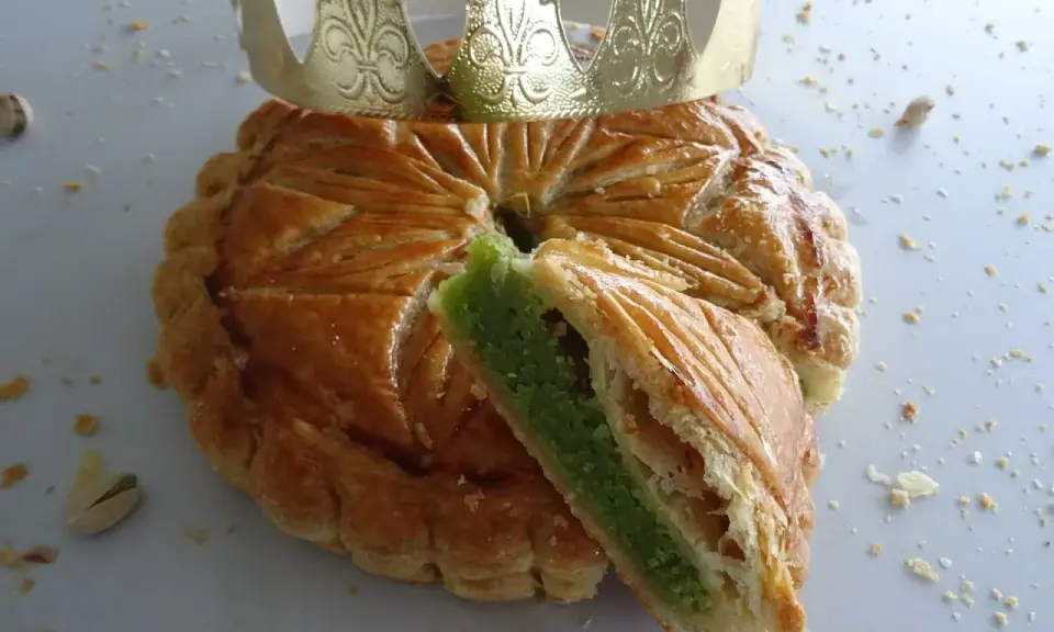 Galette des rois pistache dorée, une part coupée révèle sa garniture verte. Une couronne dorée trône sur ce délice.
