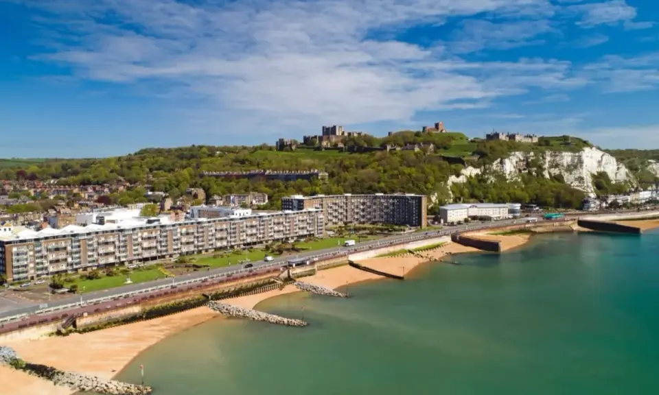 Vista aerea del porto di Dover con il castello in cima alla scogliera bianca. Edifici moderni e spiagge sabbiose si affacciano sul mare turchese.