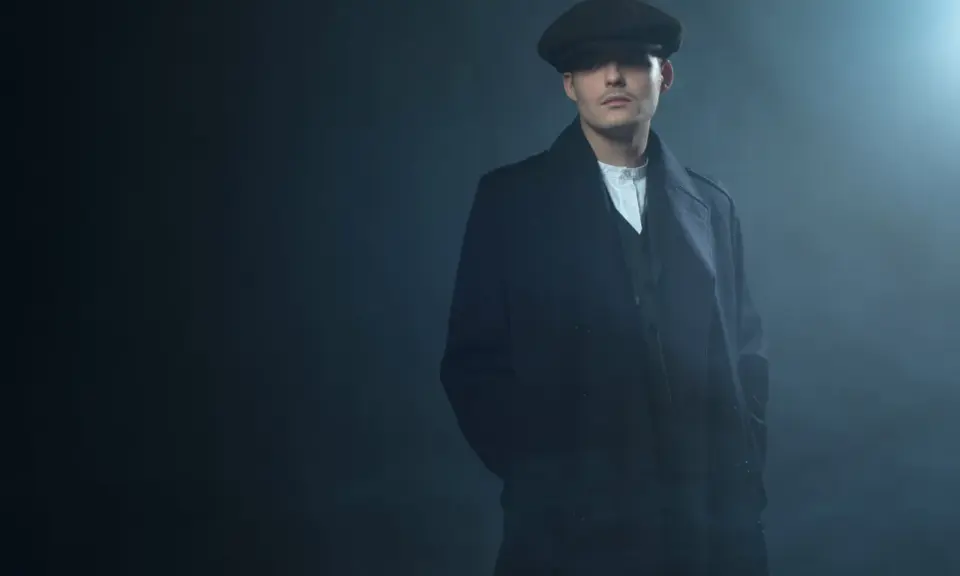 Młody mężczyzna w czapce i płaszczu, z charakterystyczną fryzurą w stylu Peaky Blinders, stoi w zadymionym mroku.