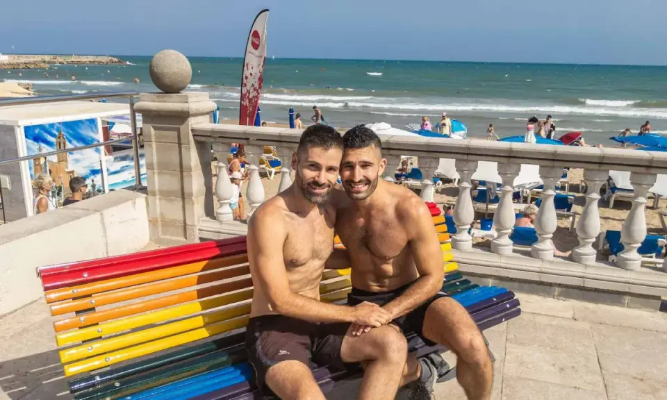 A happy couple enjoys a sunny day at the Sitges gay beach, sitting on a rainbow bench with the ocean in the background.