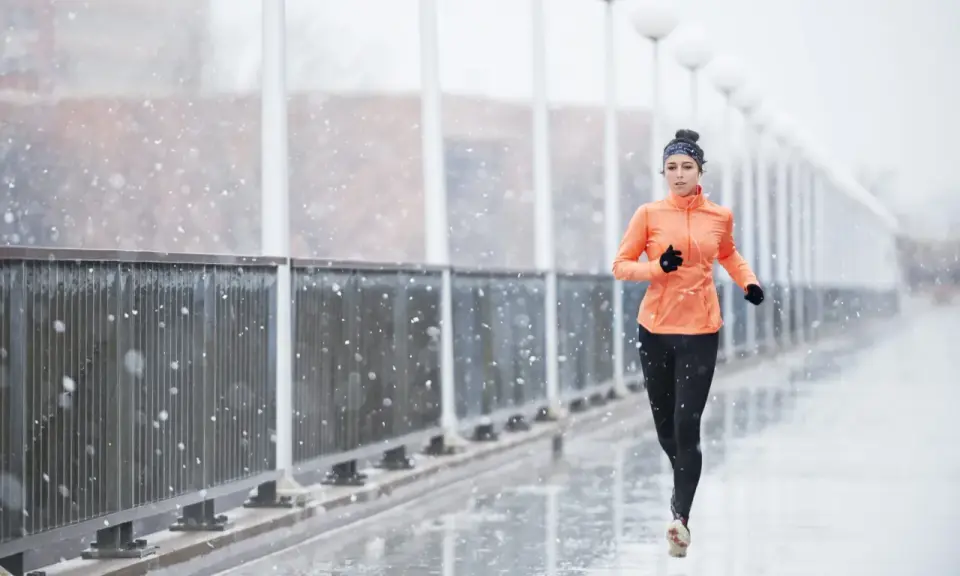 Donna corre sotto la pioggia su un ponte, con fiocchi di neve che cadono. Indossa abbigliamento sportivo arancione e nero.