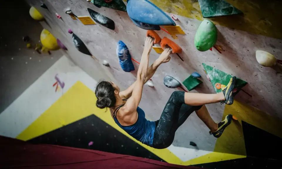 Kobieta wspina się po kolorowej ścianie, chwytając pomarańczowe uchwyty. To właśnie bouldering – sportowa wspinaczka bez liny.