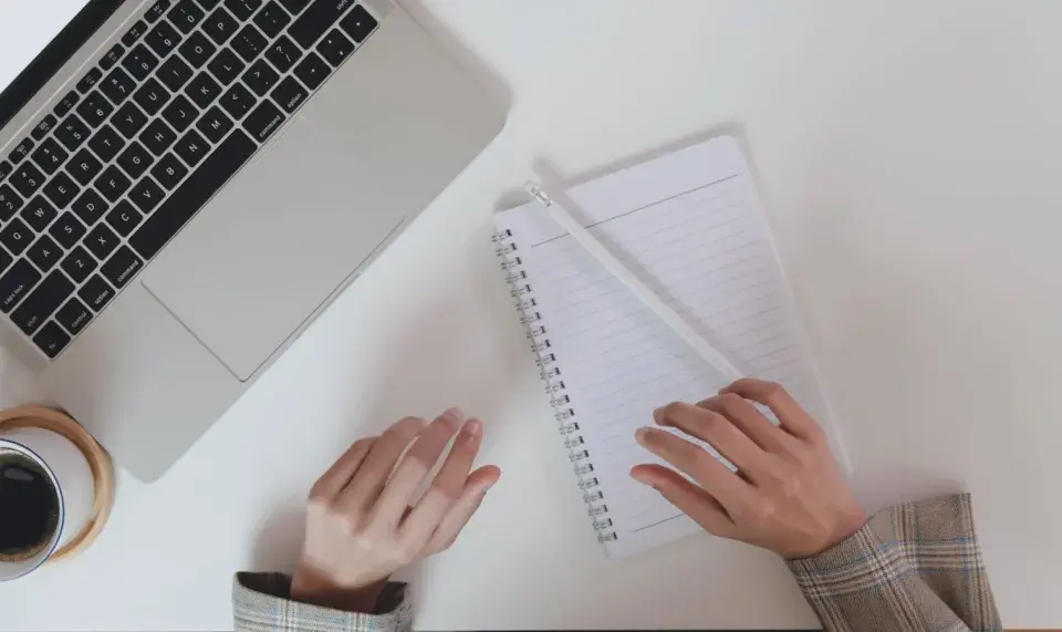 Ręce piszą notatki do CV po niemiecku przy laptopie, obok filiżanka kawy.