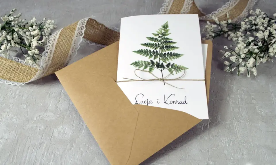 Eleganckie zaproszenie ślubne z paprocią i imionami pary. W zestawie koperta i ozdobna wstążka. To, co musi zawierać zaproszenie, to jego piękny design.