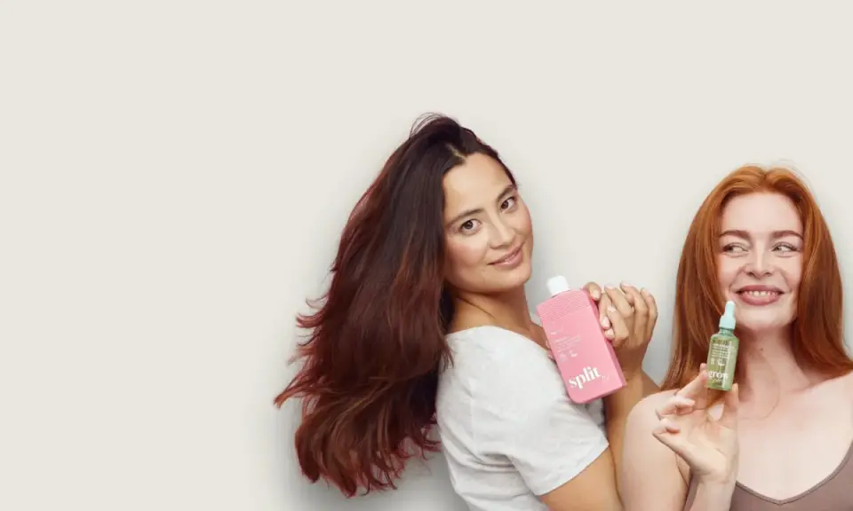 Kobiety prezentują kosmetyki do włosów. Jedna trzyma różową butelkę, druga małą buteleczkę z serum, które zawiera olejek jojoba właściwości odżywcze.