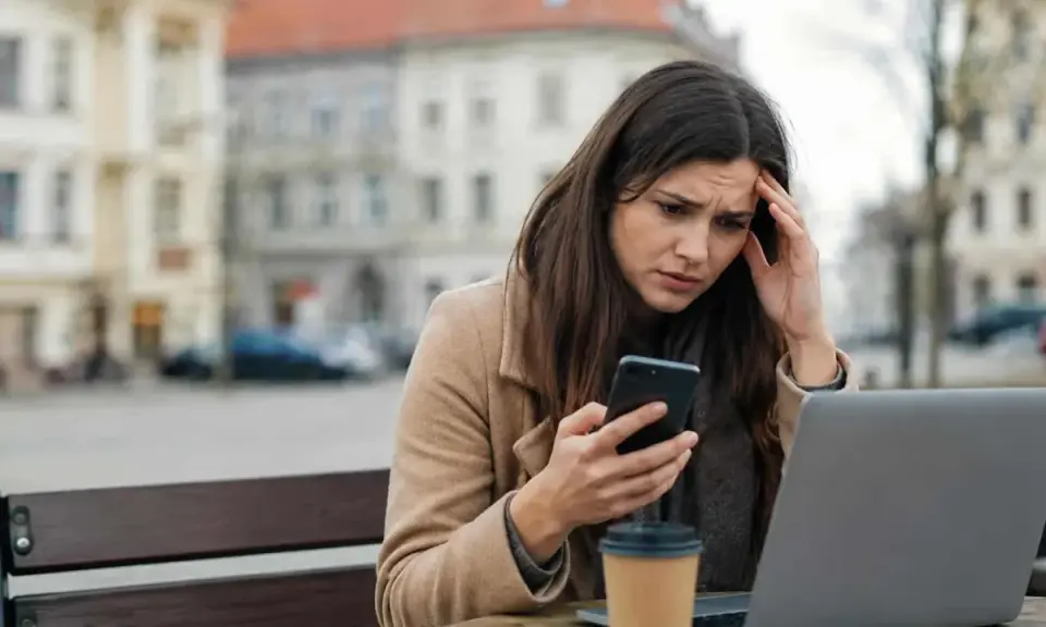 Zmartwiona kobieta czyta na telefonie coś, co wygląda na zniesławienie. Trzyma się za głowę, siedząc przy stoliku z laptopem i kawą.