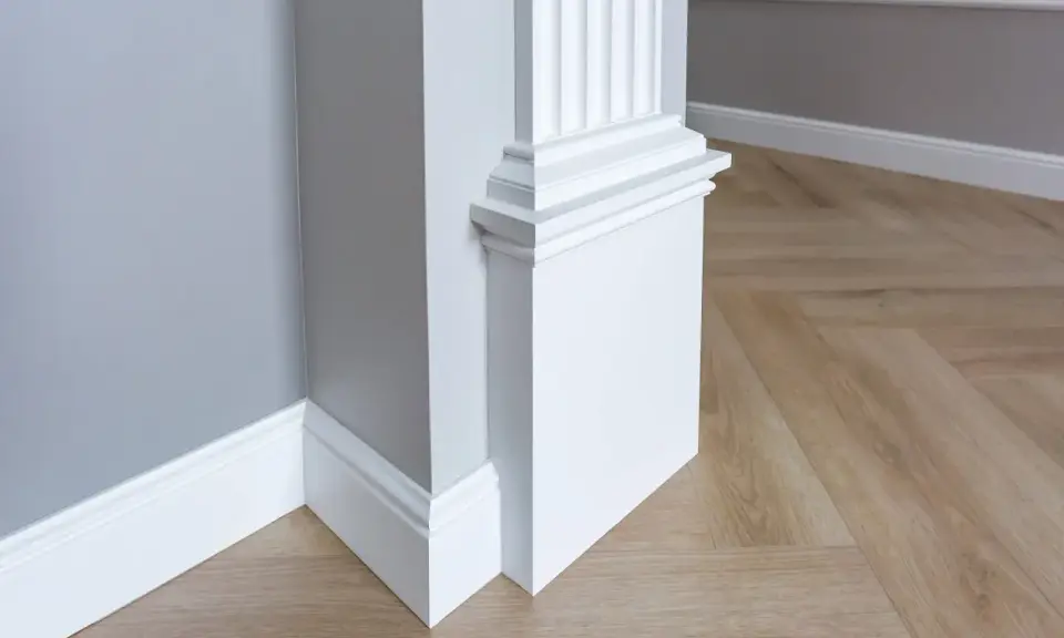 Biały pilaster z żłobieniami i elegancką bazą, idealny do wnętrzarskiego bloga. Drewniana podłoga w jodełkę i szara ściana tworzą stylowe tło.